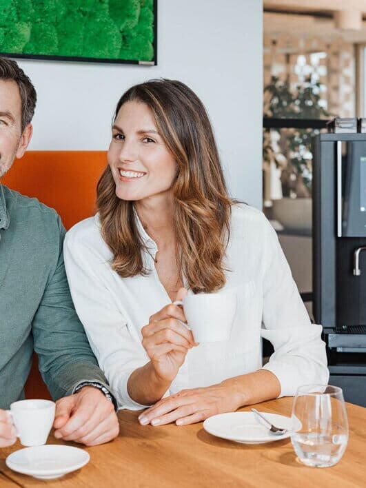 Lächelnde Frau und Mann genießen gemeinsam Kaffee im Büro, zubereitet mit einem Kaffeevollautomaten der BARISTA-Serie von Kaffee Partner.
