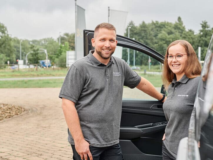 Zwei Servicemitarbeitende stehen vor einem Firmenfahrzeug mit offener Tür und lächeln in die Kamera. Sie tragen Arbeitskleidung mit Firmenlogo.