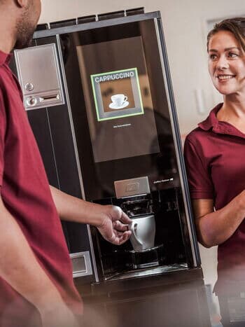 Mitarbeitende stehen an der Kaffeemaschine, waehrend frischer Cappuccino zubereitet wird.