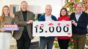 Fünf Personen stehen vor einem weihnachtlich geschmückten Hintergrund mit einem Weihnachtsbaum. Sie halten ein Schild mit der Aufschrift „15.000“, auf dem kleine Weihnachtsmützen dekorativ angebracht sind.