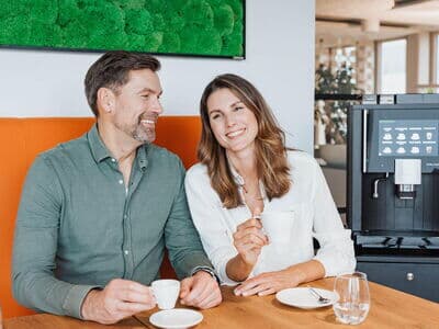 Man and woman drinking coffee next to a fully automatic coffee machine while sitting next to each other at a table