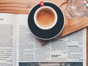 Eine Zeitung liegt auf einem Tisch, darauf steht eine Tassee Kaffee und ein Glas Wasser.