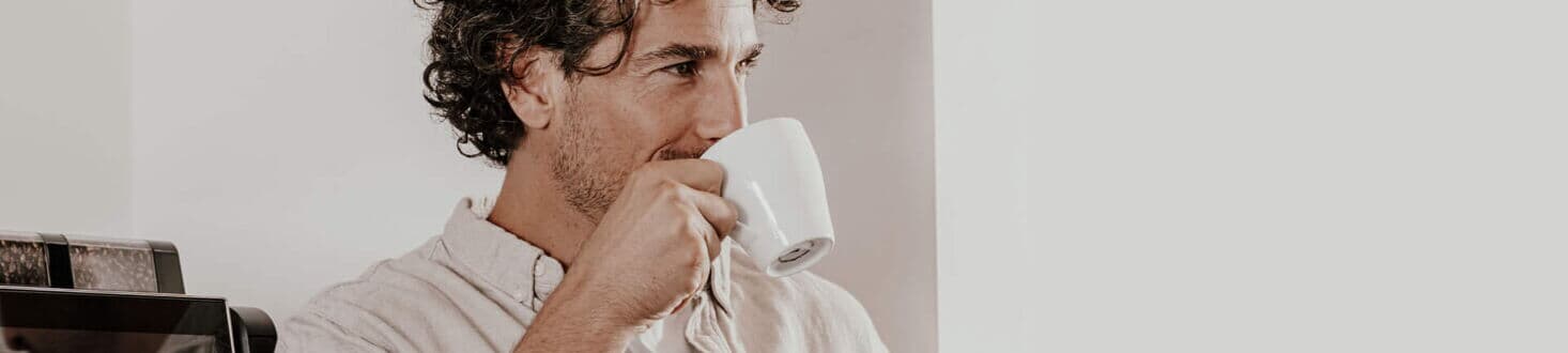 Ein Mann mit lockigem Haar genießt eine Tasse Kaffee, während er neben einem Kaffeevollautomaten von Kaffee Partner steht.